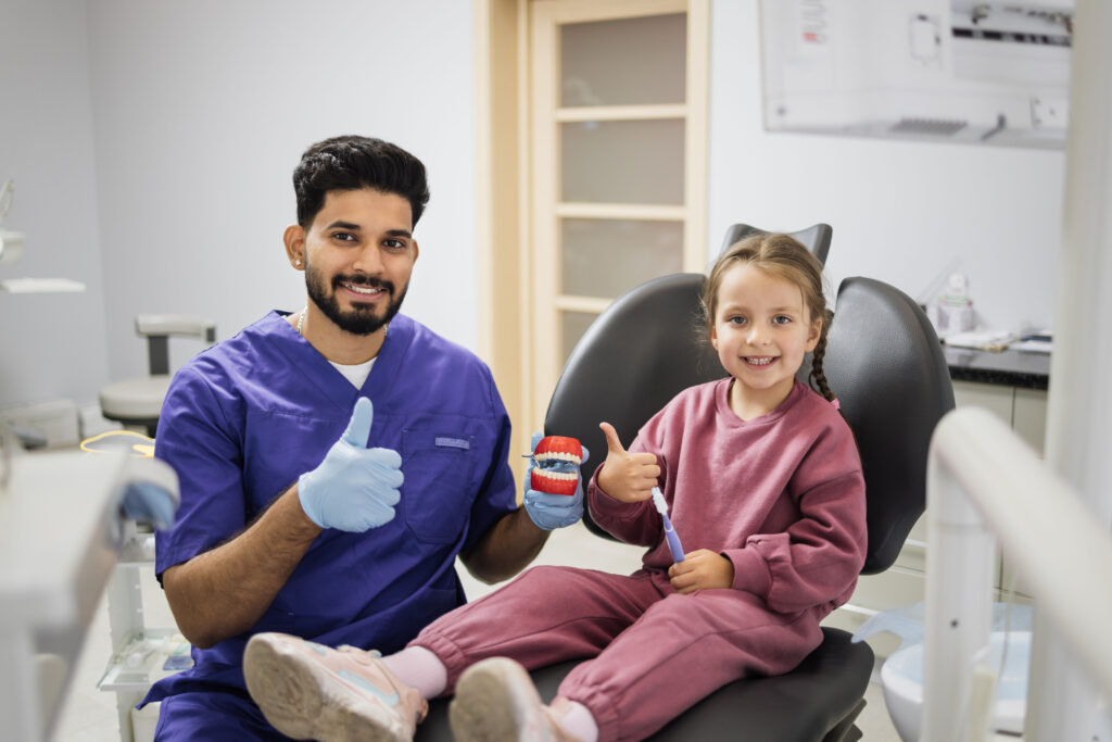 Lakeside smile Happy confident male dentist tells little child girl how to brush the teeth on artificial jaw model showing thumbs up. Caries prevention, pediatric dentistry, milk teeth hygiene concept.