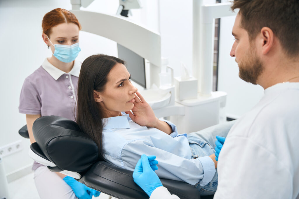 Lakeside smile Woman patient complaining to dentist on dull and constant toothache sitting in orthodontic chair, stomatologist and nurse attentively listening to client before oral cavity examination